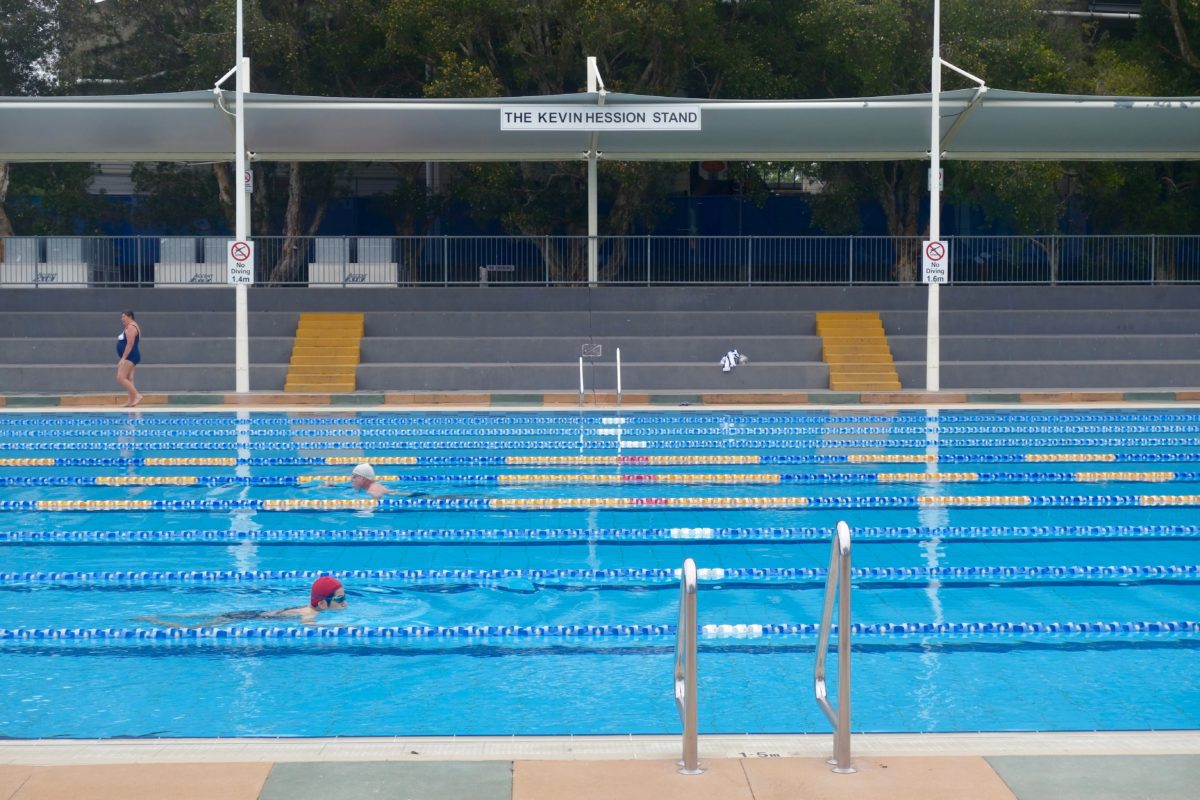 The old pool’s grey concrete grandstand with yellow steps under a shade sail signed The Kevin Hession Stand, with swimmers doing laps in the foreground.