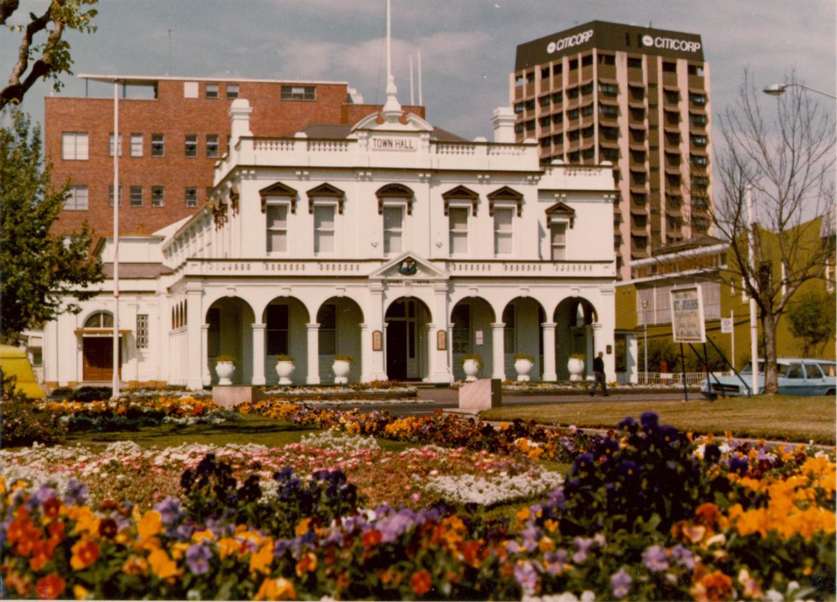 Parramatta Town Hall, a white two-storey colonnaded building, behind colourful flower beds, with an office tower behind it.