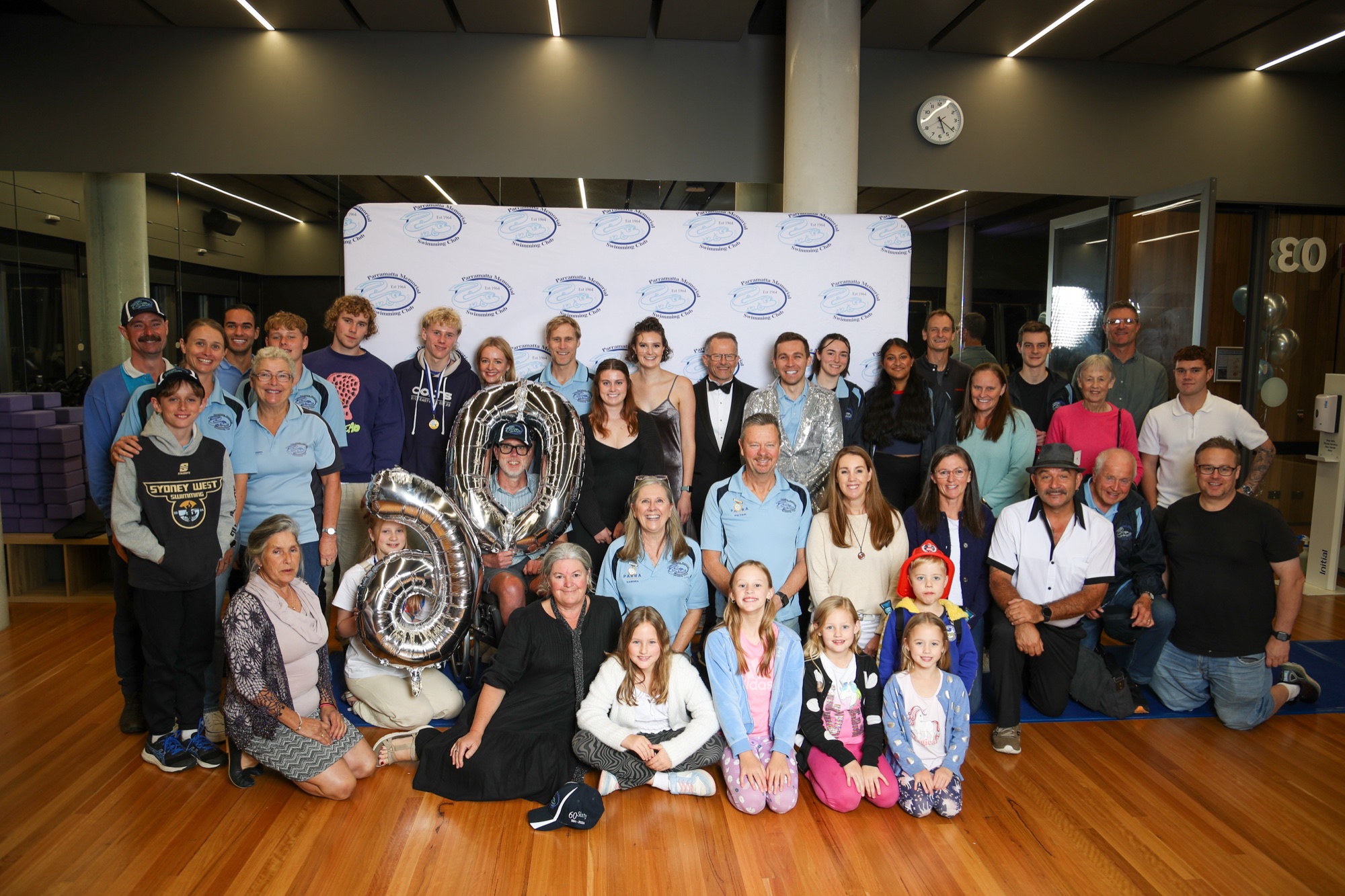 A large group of Club members of all ages, many in light-blue Club shirts, gathered in front of a PMSC logo backdrop at Parramatta Aquatic Centre, with silver “60” balloons at the front.