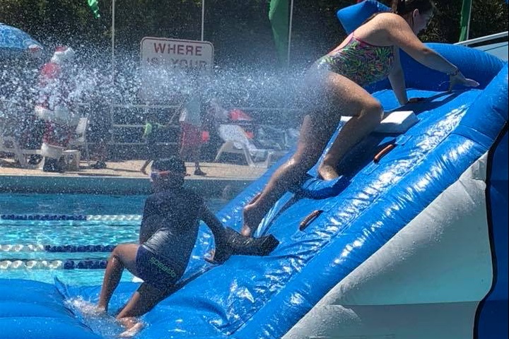 Swimmers climbing a large blue inflatable in the pool while water sprays across it.