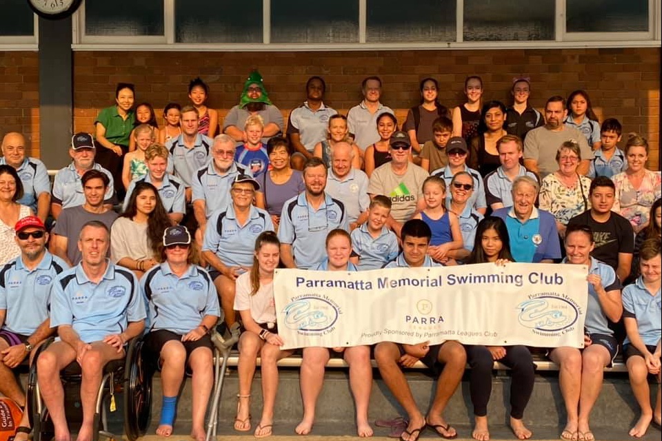 Club members of all ages, many in light-blue Club shirts, seated in the grandstand behind a Parramatta Memorial Swimming Club banner, one wearing a Christmas-tree hat.