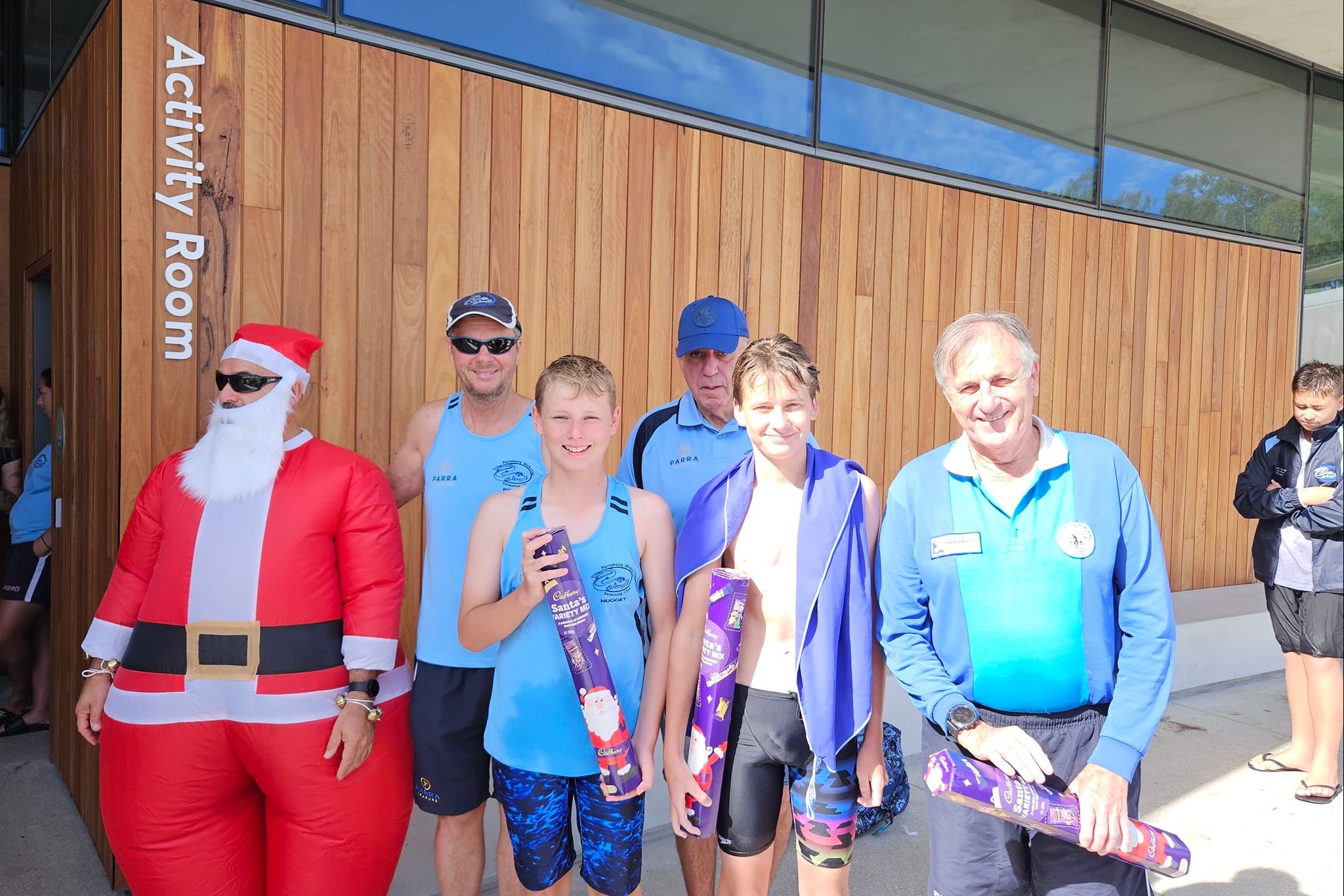 Club members holding Christmas prizes beside someone in an inflatable Santa costume, outside the Activity Room at Parramatta Aquatic Centre.