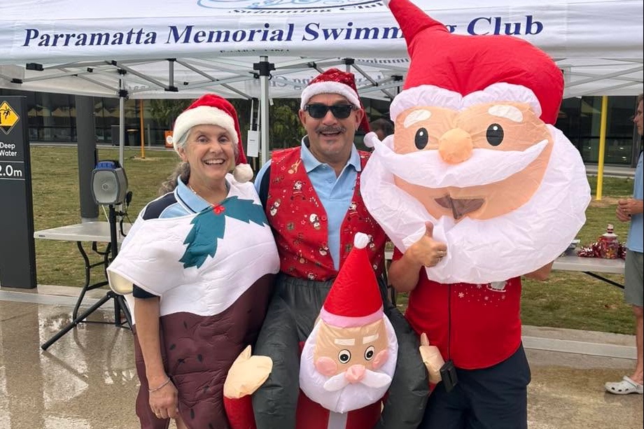 Two Club members in Christmas costumes and Santa hats with inflatable Santas, under the Parramatta Memorial Swimming Club marquee.