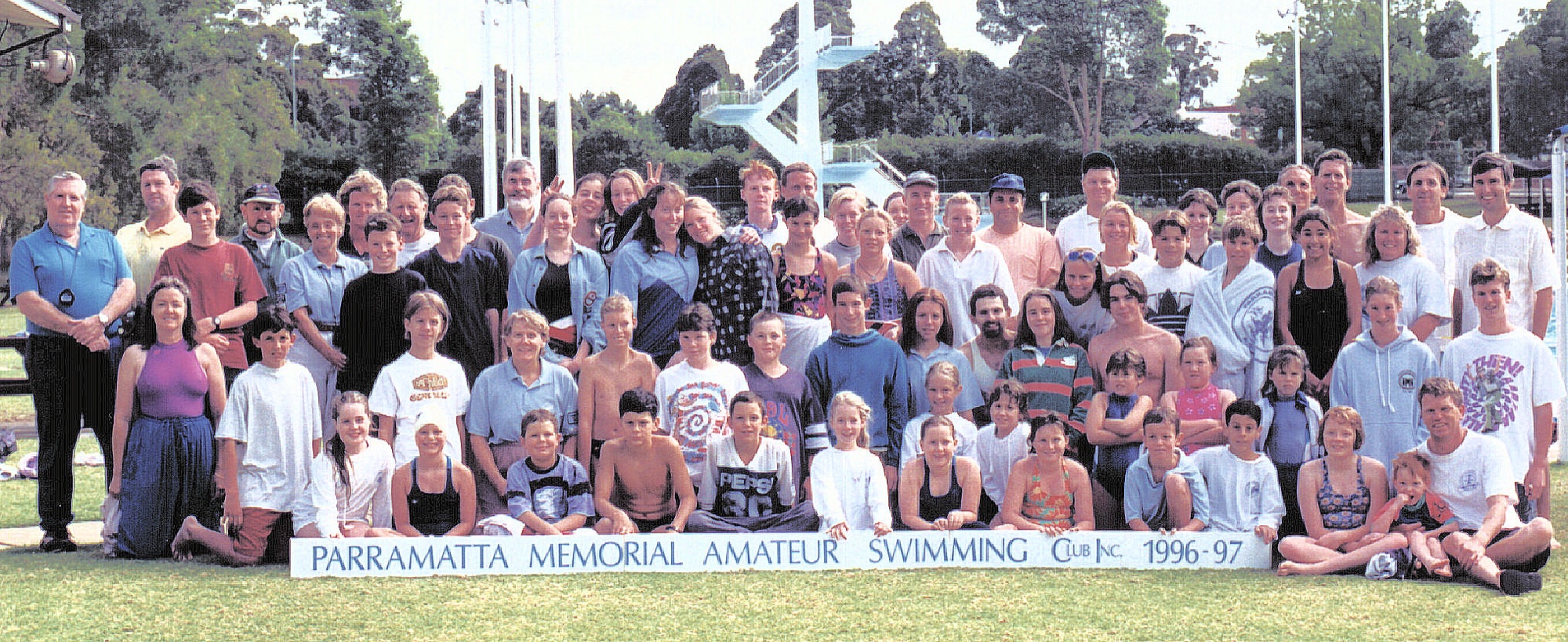 A wide group photo of Club members of all ages at the old pool, seated and standing behind a long “Parramatta Memorial Amateur Swimming Club 1996–97” banner.