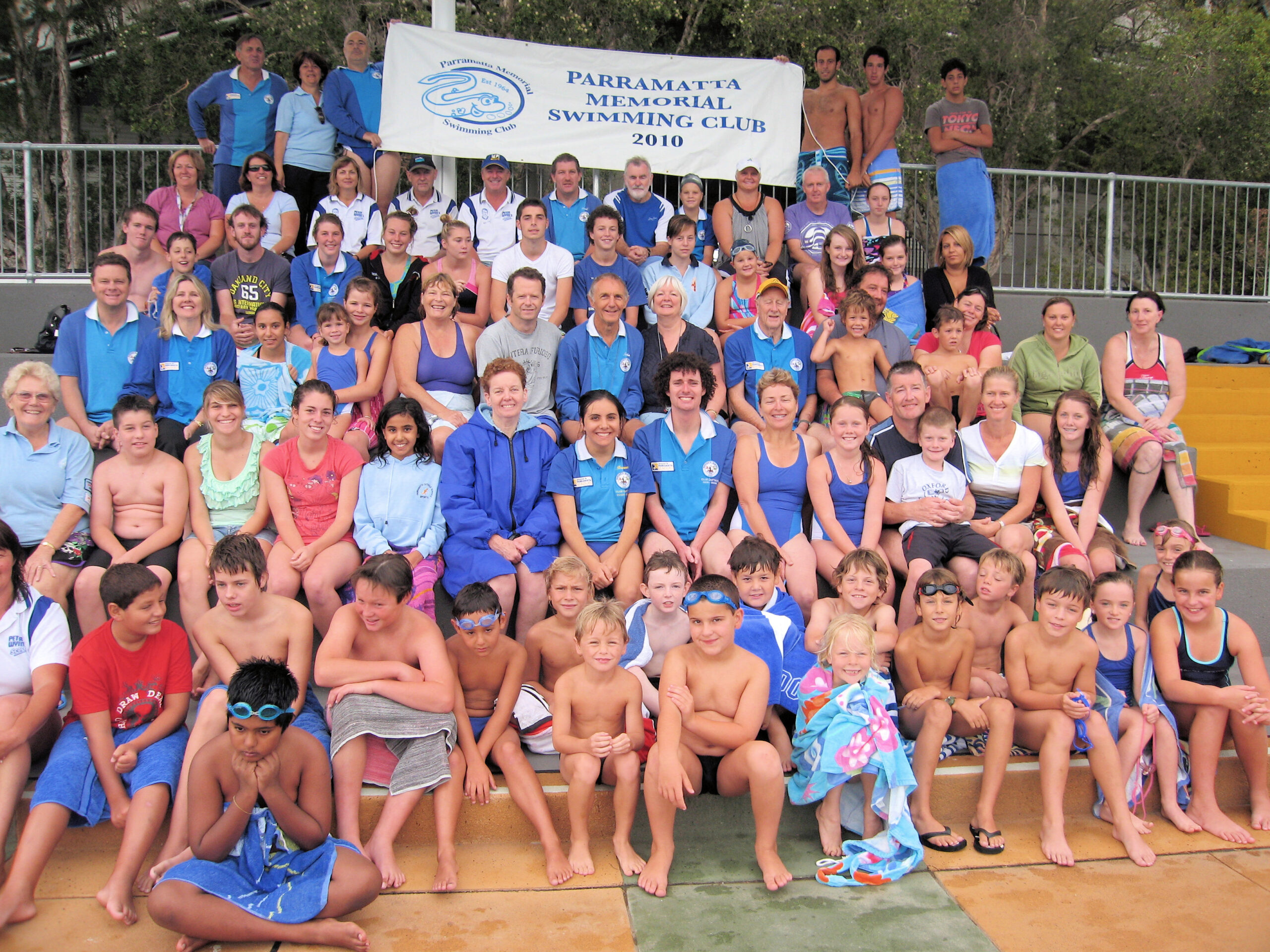Around 80 Club members of all ages seated on the grey concrete tiers of the grandstand at the old pool, beneath a banner reading Parramatta Memorial Swimming Club 2010.