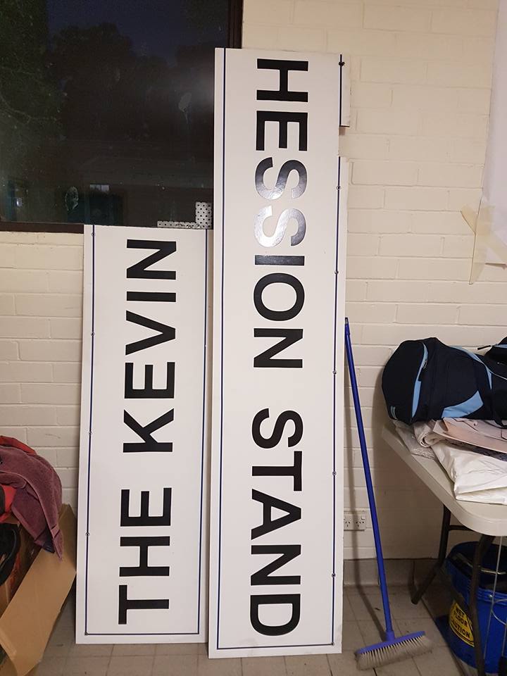 Two long white signs reading The Kevin and Hession Stand leaning against a wall indoors.