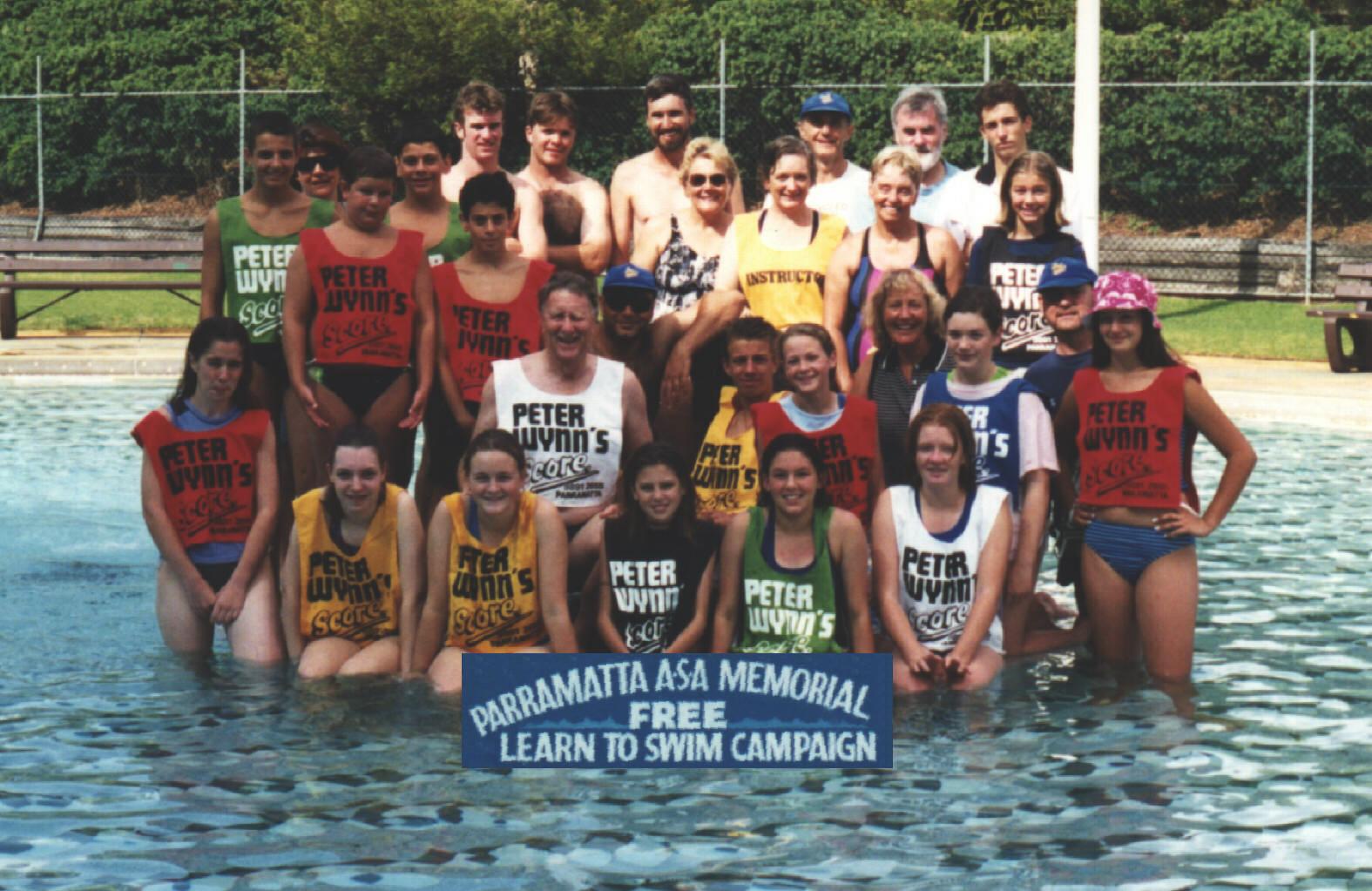 Volunteer instructors and teenage helpers in coloured bibs standing waist-deep in the old pool, behind a sign for the Parramatta ASA Memorial Free Learn to Swim Campaign.