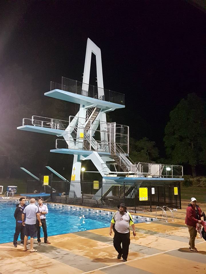 The old pool’s white diving tower floodlit at night, with people on the pool deck below.