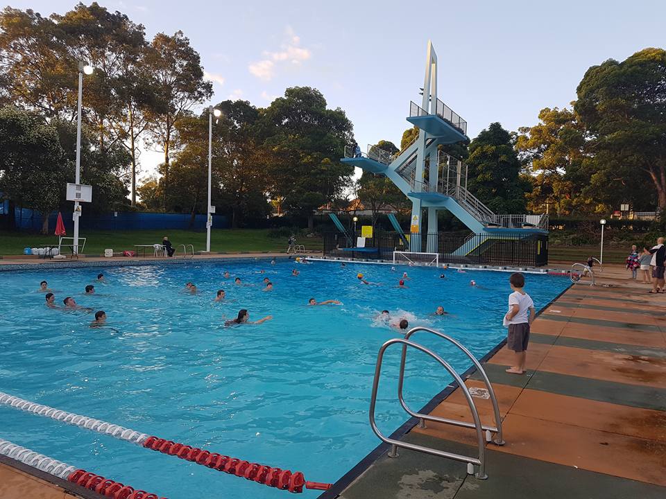 Swimmers in the old pool in evening light, with the white diving tower behind.