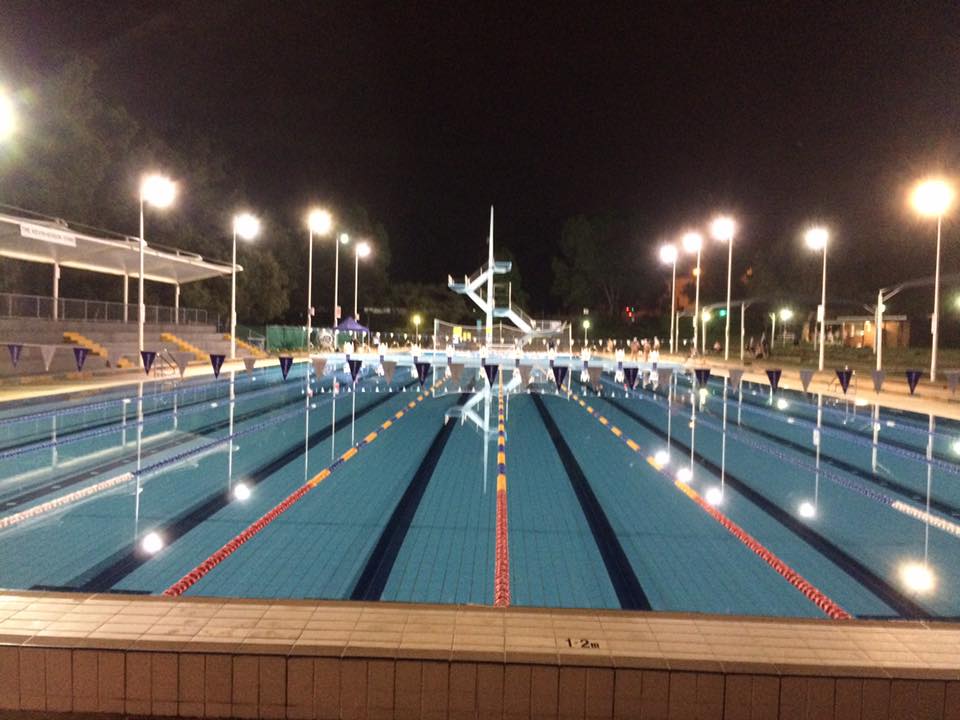 The empty 50-metre pool at night, its lanes lit by rows of lights, with the diving tower in the distance.