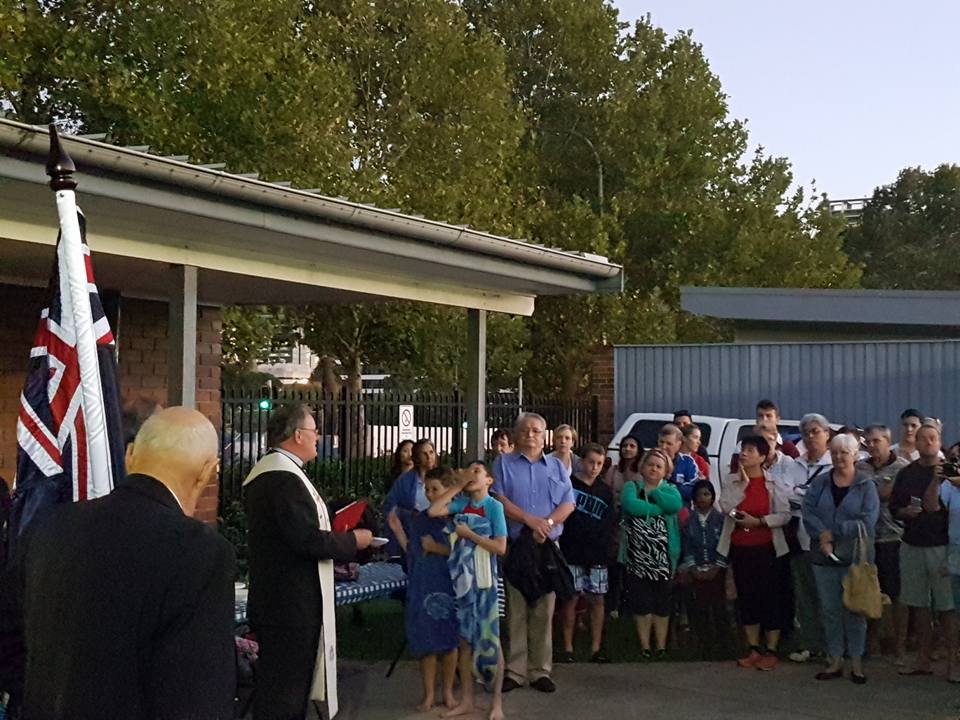 A crowd standing in silence at dusk beside the pool buildings, with an Australian flag and a priest in vestments at the front.