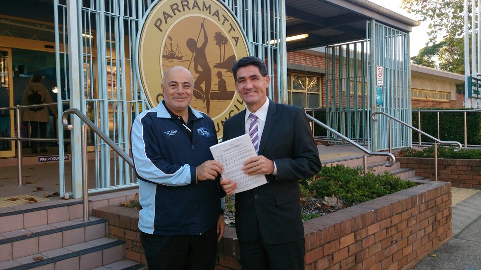 Two men in suits and Club shirts at the pool entrance, one handing a folder of papers to the other, with the Parramatta City Council emblem behind.