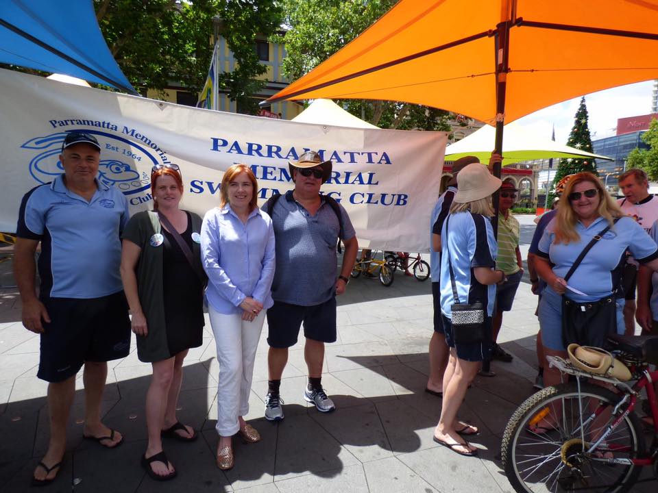 Club members in Club shirts beside the Parramatta Memorial Swimming Club banner at an outdoor rally under colourful umbrellas.
