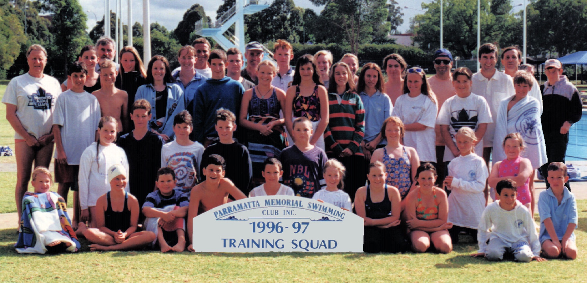 A large training squad of swimmers, parents and coaches on the grass at the old pool behind a “1996–97 Training Squad” sign, with the diving tower behind.