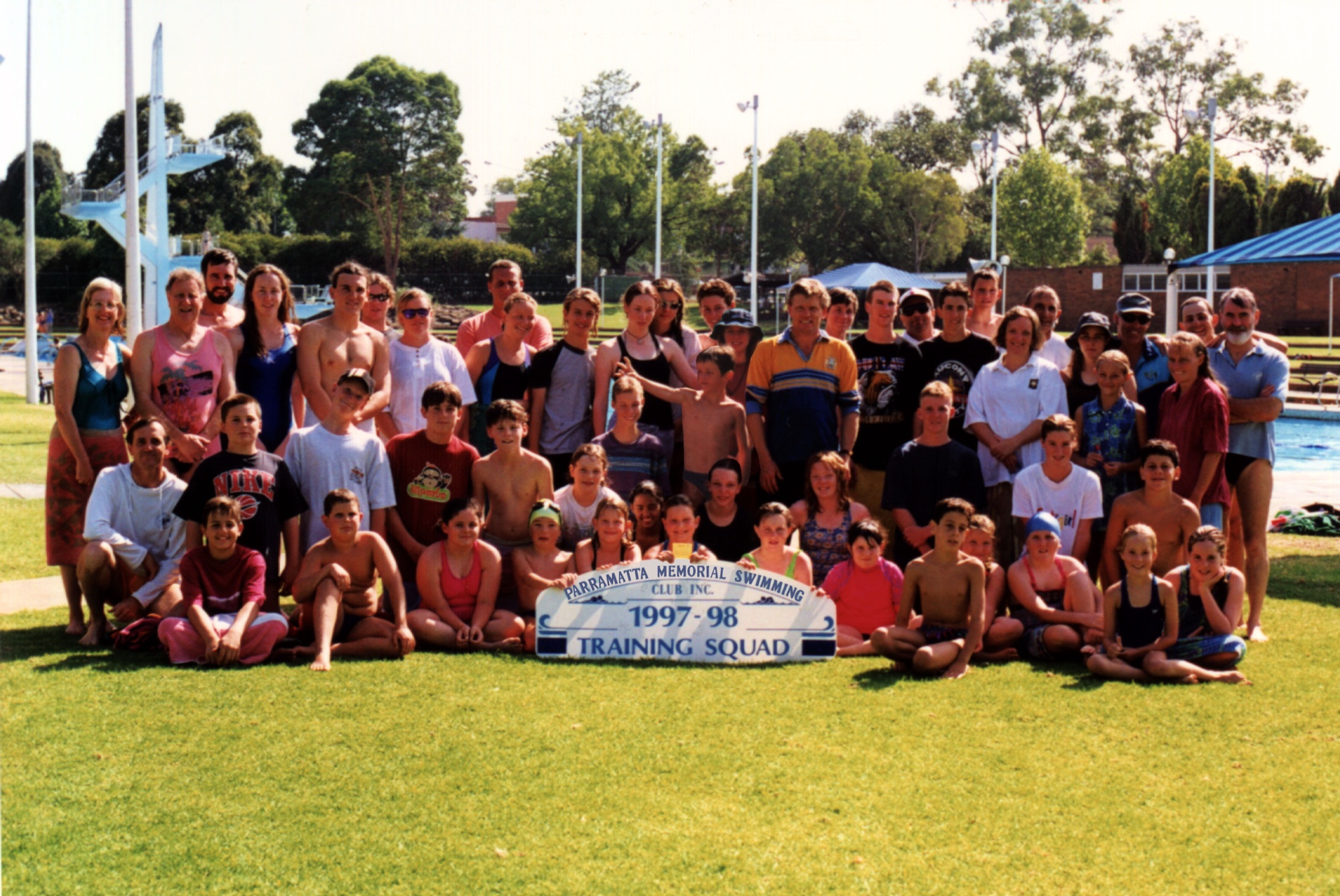 A large training squad on the lawn at the old pool behind a “1997–98 Training Squad” sign, with the diving tower and grandstand shade behind.