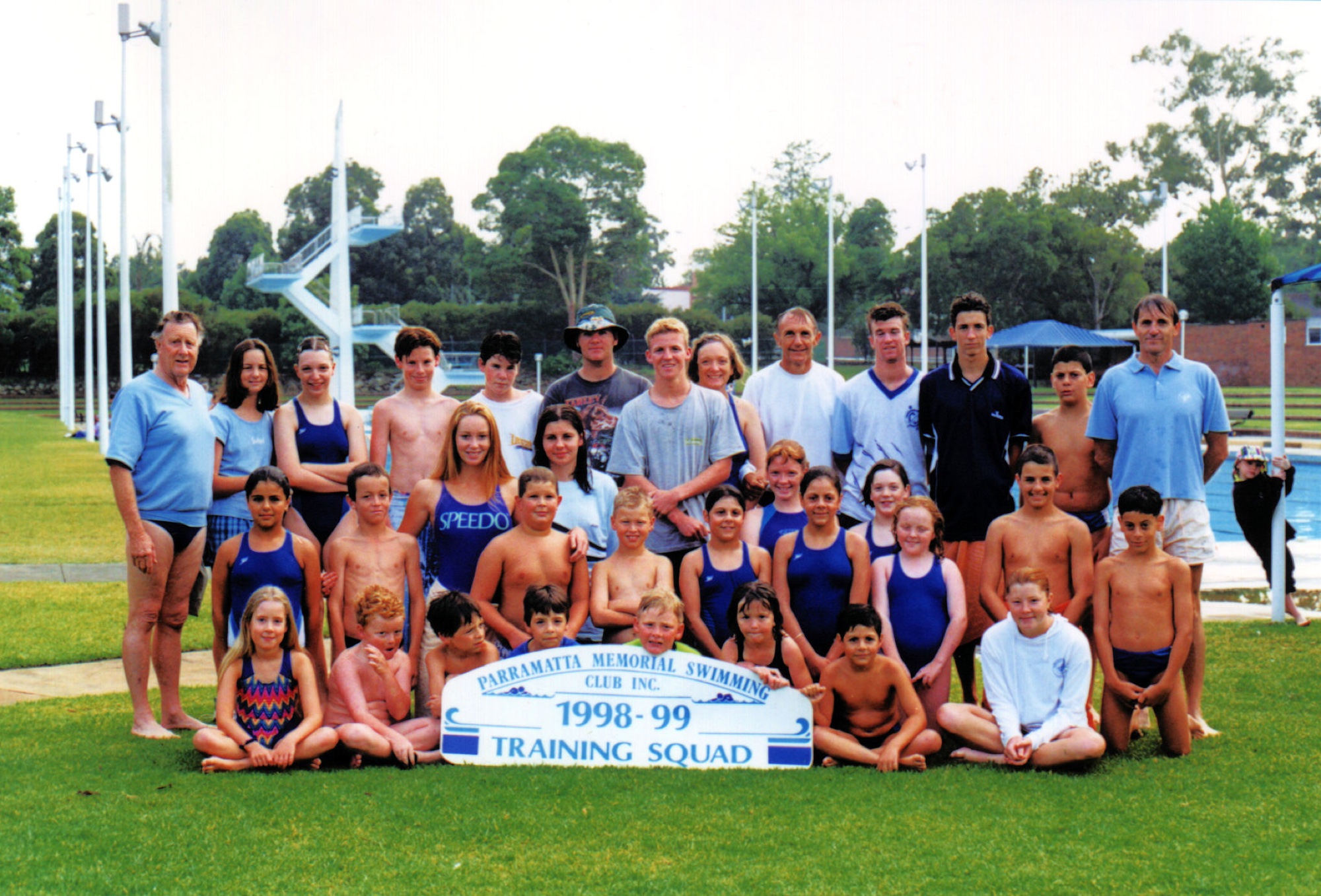 Swimmers in swimsuits and coaches on the lawn at the old pool behind a “1998–99 Training Squad” sign, with the diving tower behind.